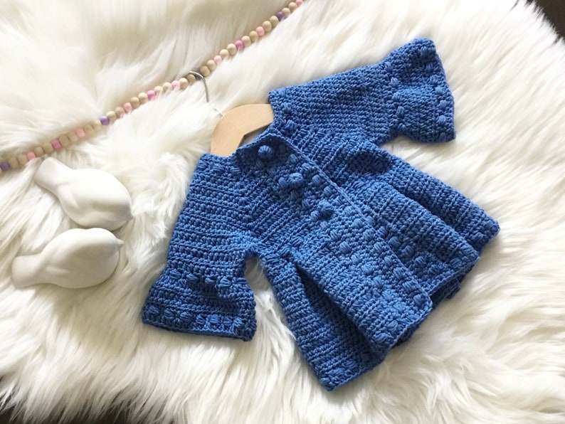 May include: A blue crocheted baby cardigan with a button closure and ruffled sleeves. The cardigan is laid out on a white fluffy surface.