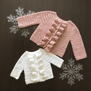 Może przedstawiać: Dwa dzianinowe kardigany dla niemowląt, jeden w kolorze różowym, a drugi w białym, z falbanką na przodzie. Oba kardigany mają długie rękawy i zapięcie na guziki.