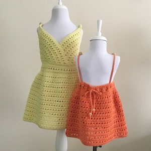 May include: Two crocheted dresses, one yellow and one orange, on mannequins. The dresses have a simple, open-stitch design.