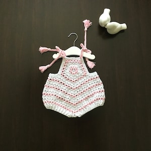 May include: White crochet baby romper with pink trim and a pink crocheted flower design. The romper has straps with pink tassels.