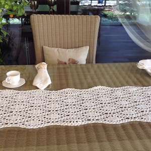 May include: A white crocheted table runner with a delicate pattern lays on a brown wicker table. A white teacup and saucer are on the table to the left of the runner. A white ceramic dish is on the table to the right of the runner.