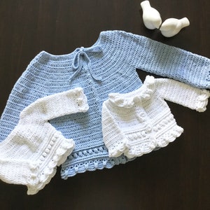 May include: Two crocheted baby sweaters, one in light blue and one in white. The sweaters have a delicate crocheted trim and are laid out on a dark brown surface.
