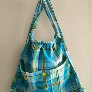 Puede incluir: Una bolsa de cordón de cuadros azules y verdes con un botón amarillo. La bolsa está colgada de un gancho en una pared blanca.