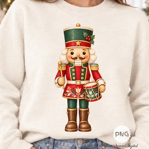 Op de afbeelding: Een crèmekleurig sweatshirt met ronde hals en een centrale afbeelding van een notenkraker-soldaat. De notenkraker draagt een groene hoed met hulst, een rood jasje met gouden randen en een groene broek. Hij houdt een trommel en een stok vast.