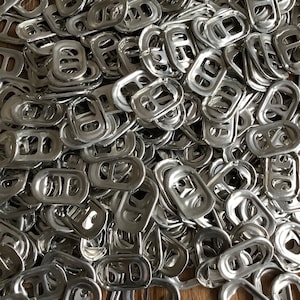 Könnte beinhalten: Nahaufnahme eines Haufens silberner Aluminium-Dosenlaschen. Die Laschen sind oval geformt mit einer rechteckigen Öffnung und einer kleinen Schlaufe. Die Laschen sind auf einer Holzoberfläche verstreut.