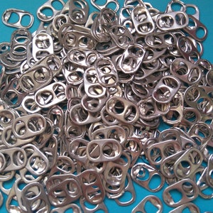 Op de afbeelding: Een grote stapel zilveren ringtrekkers van aluminium drankblikjes. De ringtrekkers zijn willekeurig gerangschikt op een blauwe achtergrond.
