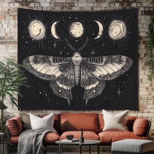May include: Black tapestry featuring a detailed moth illustration with celestial elements. The design includes phases of the moon and stars. The moth has intricate wing patterns in shades of cream and gray. The tapestry is displayed above a red sofa.