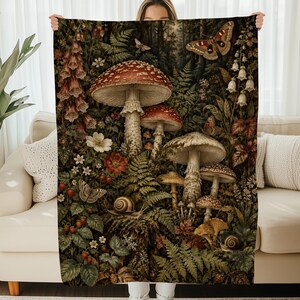 May include: A rectangular throw blanket featuring a detailed forest floor illustration with large red and white spotted mushrooms, various green ferns, small white flowers, pink foxgloves, brown snails, and a large moth with patterned wings.