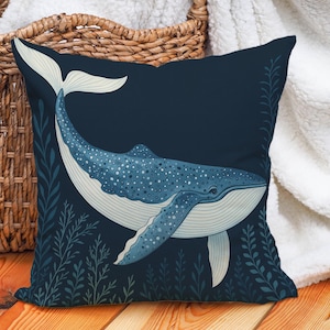May include: Square cushion with a blue whale design. The whale is illustrated with white and blue details, set against a dark blue backdrop with aquatic plants. The cushion is near a wicker basket and a white blanket. The design evokes a nautical theme.