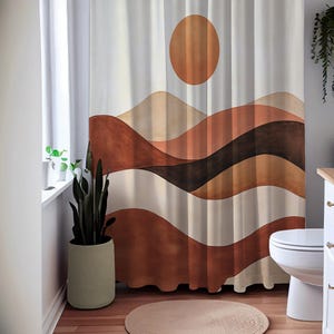 May include: A white shower curtain with a landscape design featuring a large orange sun and layered brown and beige wavy lines. The curtain is hanging in a bathroom with a toilet, a round rug, and a potted plant.