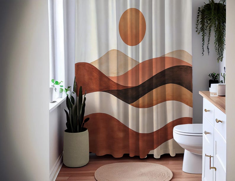 May include: A white shower curtain with a landscape design featuring a large orange sun and layered brown and beige wavy lines. The curtain is hanging in a bathroom with a toilet, a round rug, and a potted plant.