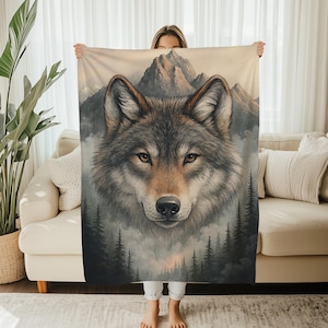 May include: A blanket with a detailed illustration of a wolf's face, set against a backdrop of mountains and a forest. The wolf has brown eyes and grey and brown fur. The blanket is held up by a person.