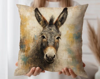 May include: Square decorative pillow featuring a detailed painting of a donkey's face. The artwork uses a muted color palette with shades of gray, brown, and yellow. The pillow is held by a person.