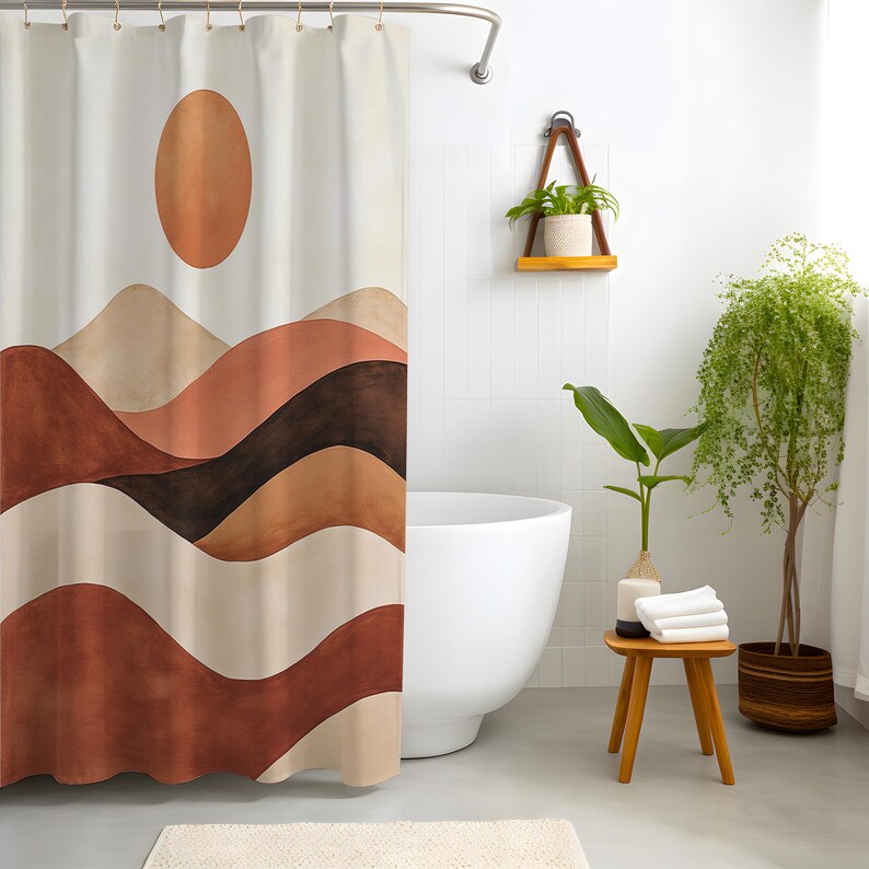 May include: A beige shower curtain featuring a watercolour design of a sun and layered mountain range in shades of brown, tan, and rust. The curtain hangs in front of a white bathtub. A small wooden stool holds towels and a vase with a plant.