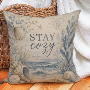 May include: Square decorative pillow with a coastal theme. The pillow features a vintage design with seashells, starfish, and blue and beige accents. The text "STAY COZY" is displayed on the front, along with the phrase "the ocean is my happy place."