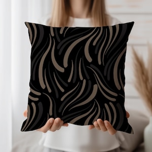 May include: Square decorative pillow with a black background and a repeating pattern of flowing, abstract shapes in shades of brown and gray. The pillow is held up by a person.