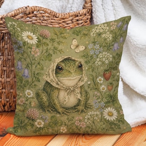 Könnte beinhalten: Ein quadratisches Zierkissen mit einem grünen Frosch, der ein gemustertes Kopftuch trägt, inmitten einer detaillierten Illustration von Wildblumen, Klee, Erdbeeren und einem Schmetterling auf olivgrünem Hintergrund mit weicher Stoffstruktur.
