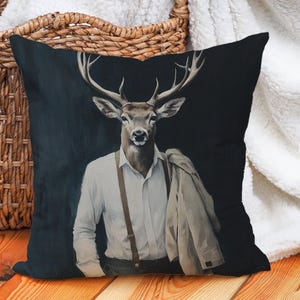 May include: Square cushion with a deer illustration wearing a white shirt, braces, and a jacket. The cushion has a dark background. It is placed on a wooden floor, near a wicker basket and a white blanket.