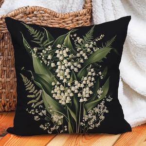 Peut inclure: Un coussin noir avec un motif floral représentant des fleurs blanches de muguet et des fougères vertes.