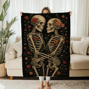 Op de afbeelding: Een zwarte deken met een romantische illustratie van twee omarmende skeletten, versierd met kleurrijke bloemen. De skeletten hebben bloemen in hun haar en zijn omringd door rode, oranje en blauwe bloemaccenten. De deken wordt door een persoon vastgehouden.