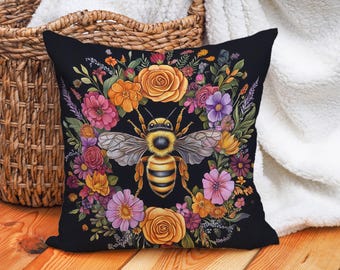 May include: Black square throw pillow featuring a detailed illustration of a bee surrounded by a floral wreath. The bee is yellow and black with detailed wings. The flowers are in shades of pink, orange, yellow, and purple.