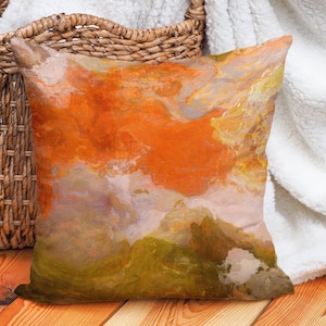 May include: Square decorative pillow with an abstract design. The pillow features a blend of orange, yellow, green, and white hues, creating a textured, artistic appearance. The pillow is placed on a wooden surface.