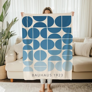 Op de afbeelding: Een rechthoekige deken met een geometrisch ontwerp van blauwe halve cirkels en cirkels op een crèmekleurige achtergrond. De tekst "BAUHAUS 1923" staat onderaan gedrukt. De deken wordt door een persoon vastgehouden.