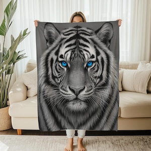 Puede incluir: Una manta de tigre gris y blanco con llamativos ojos azules. La manta presenta una ilustración detallada de la cara de un tigre, con rayas negras y una textura suave y peluda. El diseño está centrado en un fondo gris.