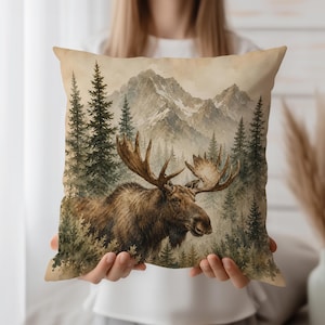 May include: A square throw pillow featuring a watercolour-style illustration of a brown moose with large antlers standing in a forest of evergreen trees. In the background, tall, snow-capped mountains rise against a soft, muted beige sky.