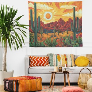 Puede incluir: Un tapiz que presenta un paisaje des&eacute;rtico con una puesta de sol naranja brillante, monta&ntilde;as y cactus. El tapiz est&aacute; colgado en una pared blanca sobre un sof&aacute; gris con almohadas coloridas y una mesa de caf&eacute;.