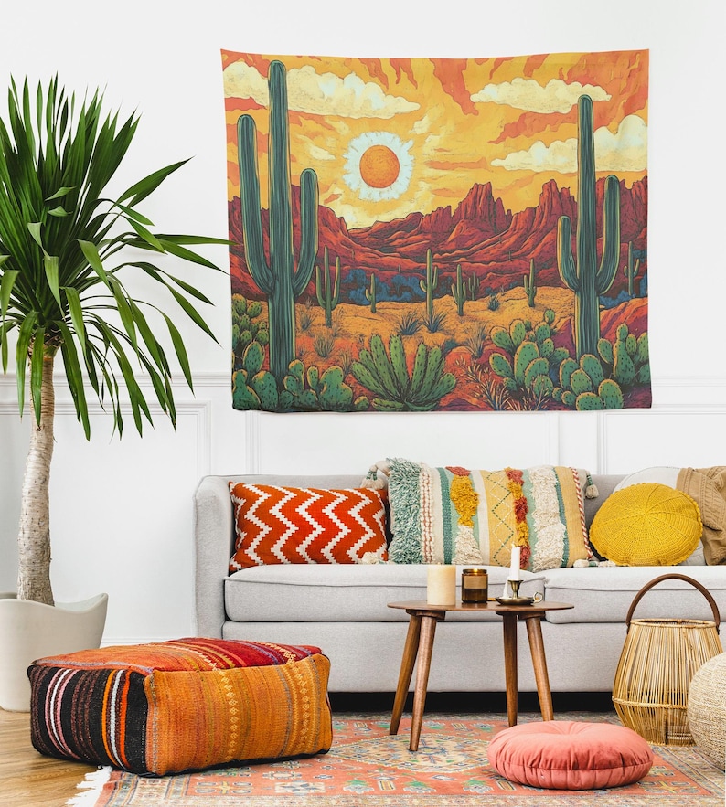 Puede incluir: Un tapiz que presenta un paisaje des&eacute;rtico con una puesta de sol naranja brillante, monta&ntilde;as y cactus. El tapiz est&aacute; colgado en una pared blanca sobre un sof&aacute; gris con almohadas coloridas y una mesa de caf&eacute;.