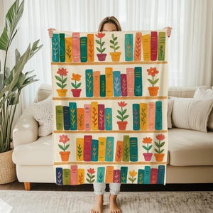 May include: A cream-coloured throw blanket with a colourful bookshelf design. The design features rows of books in various colours, potted flowers, and wooden shelves. The blanket is held up by a person.