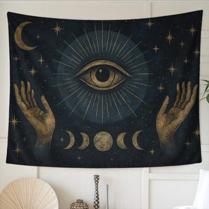 Puede incluir: Tapiz azul oscuro con un diseño central de ojo rodeado de estrellas, fases lunares y una luna creciente doradas. Manos doradas a ambos lados del ojo. El tapiz tiene una temática celestial, ideal para decoración.