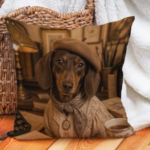 May include: A decorative square pillow featuring a dachshund wearing a brown beret, scarf, and sweater. The dog is sitting in front of a desk with books and a teacup. The pillow has a sepia tone.