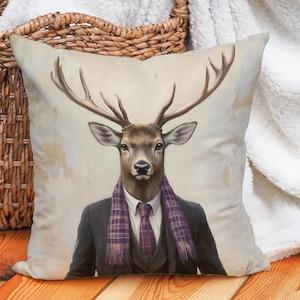 Può includere: Un cuscino decorativo con uno sfondo beige che presenta un dipinto di un cervo che indossa un abito e una sciarpa a quadri viola. Il cervo ha un manto marrone e bianco e grandi corna.