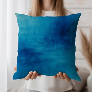 Peut inclure: Un coussin carré avec un motif abstrait bleu. Le coussin présente un dégradé de bleus, du turquoise clair au bleu marine profond, créant un effet texturé et peint. Le coussin est tenu par une personne.