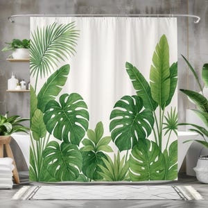 Op de afbeelding: Witte douchegordijn met een aquarelontwerp van weelderige groene tropische bladeren. De bladeren omvatten monstera, palm- en bananenbladeren. Het gordijn hangt aan een zilveren stang, wat zorgt voor een op de natuur geïnspireerde badkamerdecoratie.