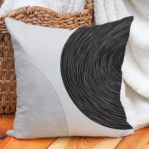 May include: A decorative cushion with a contemporary design. The cushion displays a large black, circular pattern with curved lines on the right and a light grey geometric shape on the left. It rests on a wooden surface.