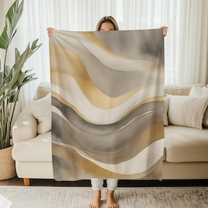 Könnte beinhalten: Eine beige-goldene Decke mit einem Aquarell-Wellenmuster. Die Decke hat fließende Linien in Gold, Beige, Weiß und Grau, die ein weiches, abstraktes Muster bilden. Die Decke wird vor einer Person gehalten.