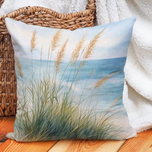 May include: Square decorative pillow with a watercolor painting of beach grass and the ocean. The pillow features shades of blue, green, and tan. The pillow is on a wooden surface.