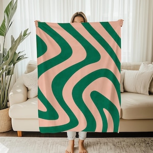 Könnte beinhalten: Eine große, weiche Decke mit Retro-Design. Die Decke hat ein sich wiederholendes Muster aus geschwungenen, dunkelgrünen Linien auf einem hellpfirsichfarbenen Hintergrund. Die Decke wird hochgehalten, um ihre Größe und ihr Design zu zeigen. Geeignet für Sofa oder Bett.