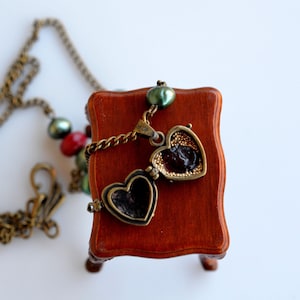Puede incluir: Un collar con un dije en forma de corazón de bronce, una cadena y cuentas verdes y rojas. El dije está abierto, revelando un compartimento más pequeño en forma de corazón en el interior.
