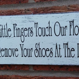 May include: White wooden sign with black text that reads "Since little fingers touch our floor, please remove your shoes at the door."