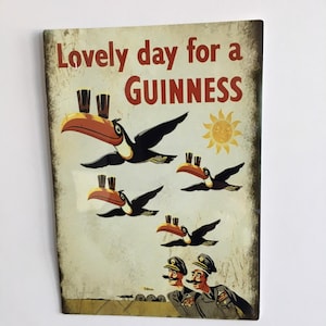 Puede incluir: Anuncio vintage de Guinness con el texto "Lovely day for a Guinness". Presenta tucanes con vasos de Guinness en sus picos, volando frente a un sol. Dos hombres uniformados con bigotes est&aacute;n en la parte inferior.