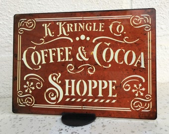 May include: A red metal sign with white lettering that reads "K. Kringle Co. Coffee & Cocoa Shoppe". The sign has a distressed look and features decorative flourishes and snowflakes.