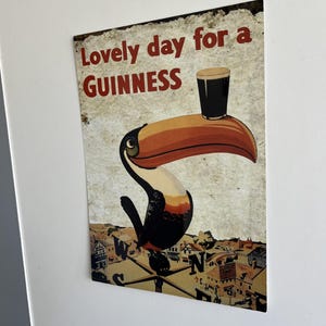 Puede incluir: Letrero met&aacute;lico vintage con un tuc&aacute;n sosteniendo una pinta de Guinness. El letrero dice "Lovely day for a Guinness" en letras rojas. El fondo muestra un paisaje urbano y una br&uacute;jula, d&aacute;ndole un toque retro.