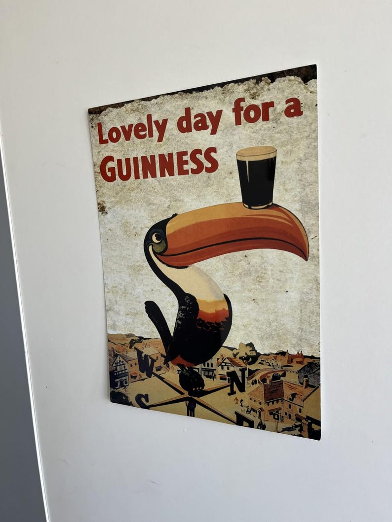 Puede incluir: Letrero met&aacute;lico vintage con un tuc&aacute;n sosteniendo una pinta de Guinness. El letrero dice "Lovely day for a Guinness" en letras rojas. El fondo muestra un paisaje urbano y una br&uacute;jula, d&aacute;ndole un toque retro.