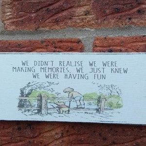 May include: White wooden sign with black lettering and a black border. The sign features a black and white illustration of two figures on a bridge. The text on the sign reads: "WE DIDN'T REALISE WE WERE MAKING MEMORIES, WE JUST KNEW WE WERE HAVING FUN."