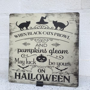 May include: A white wooden sign with black text and images. The sign says "When black cats prowl and pumpkins gleam, may luck be yours on Halloween". There are two black cats and a pumpkin with a jack-o-lantern face.