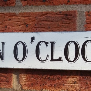 Könnte beinhalten: Ein weißes Holzschild mit schwarzer Aufschrift "GIN O'CLOCK".